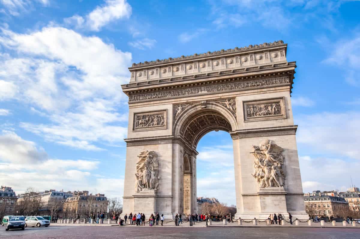 Arc de Triomphe front elevation beneath a bright cloud-filled sky with architecture details around Arc de Triomphe in Paris, France.
