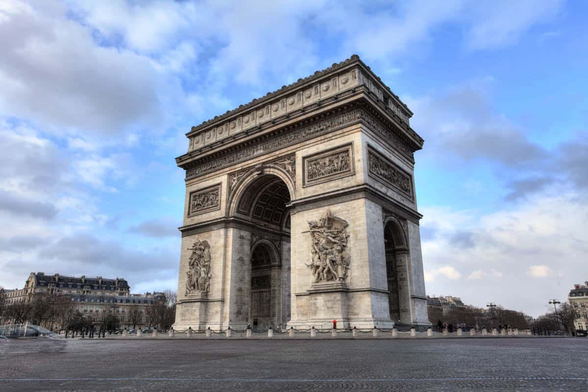 Arc de Triomphe front and side reliefs under a cloudy sky with architecture details around Arc de Triomphe in Paris, France.