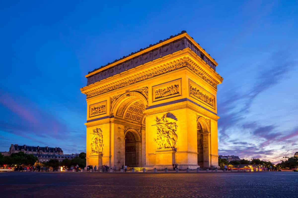 Arc de Triomphe illuminated against a deep blue evening sky with architecture details around Arc de Triomphe in Paris, France.