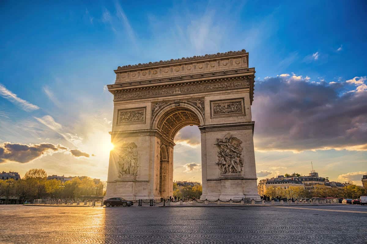Arc de Triomphe silhouetted by warm sunlight and dramatic clouds with architecture details around Arc de Triomphe in Paris, France.