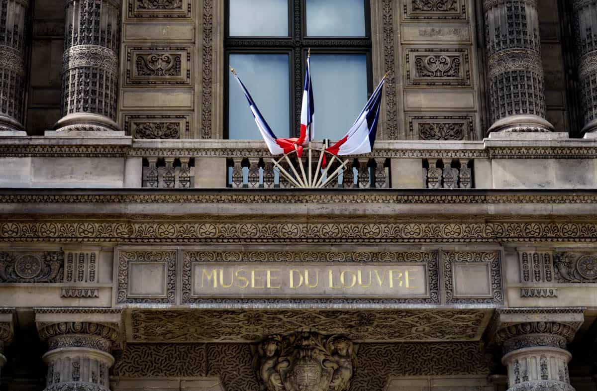 Louvre Museum in Paris, France, with Musée du Louvre inscription, French flags, and carved stone entrance detail.