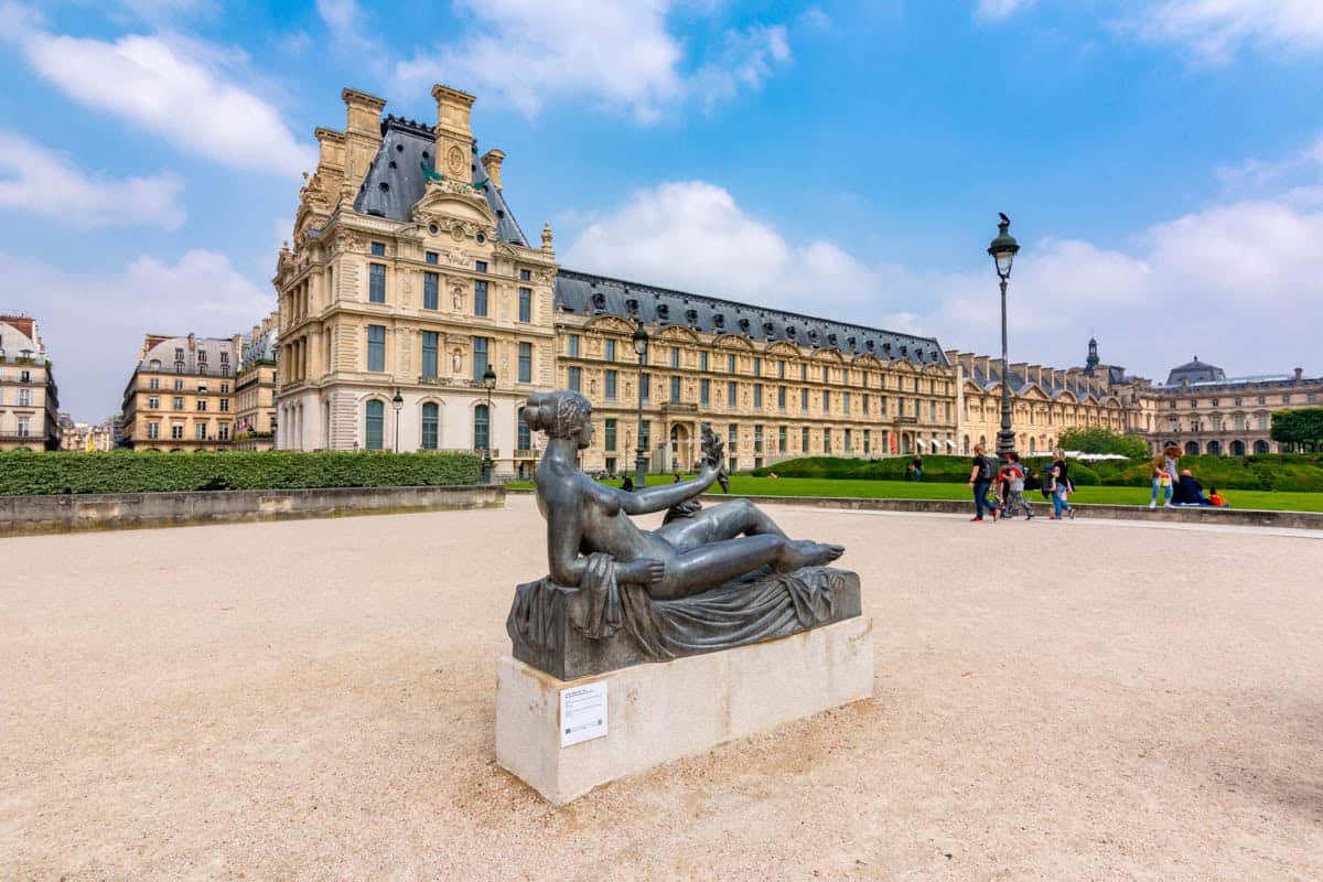 Louvre palace wing and seated stone statue across the courtyard alongside museum details at Louvre Museum in Paris, France.