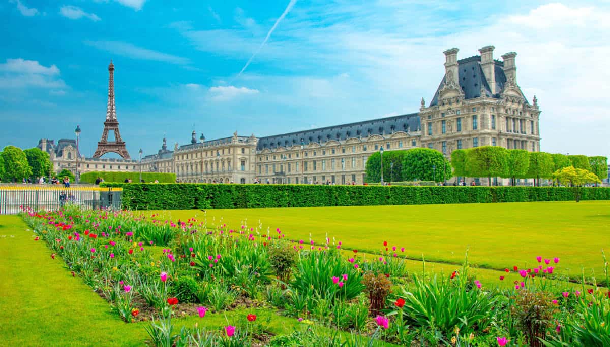 Louvre wing beyond bright Tuileries flowerbeds with the Eiffel Tower behind framed by museum and palace architecture at Louvre Museum in Paris, France.