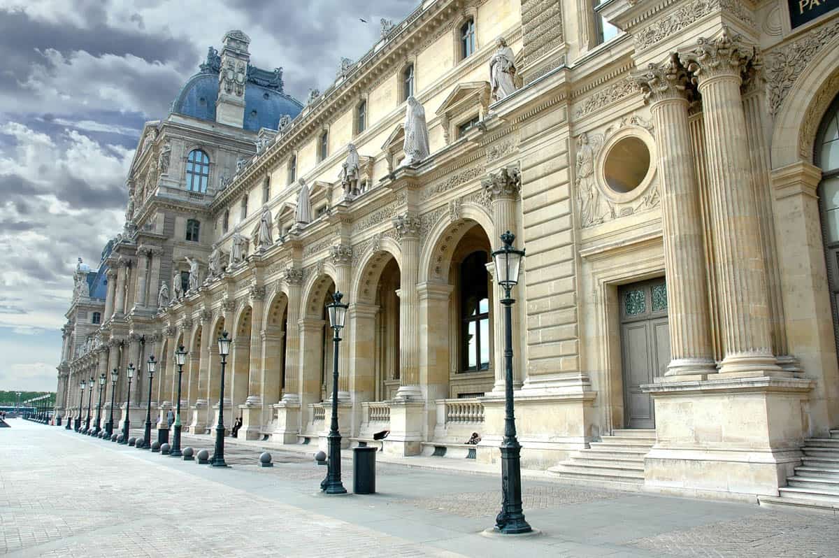 Long Louvre colonnade with arches, columns, and street lamps alongside museum details at Louvre Museum in Paris, France.