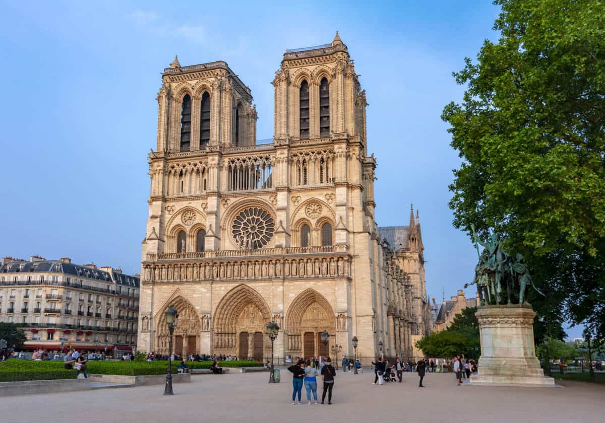 Notre-Dame west façade with trees, visitors, and a foreground statue framed by cathedral and gothic architecture at Notre-Dame Cathedral in Paris, France.