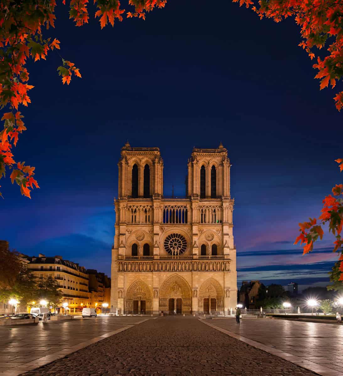 Notre-Dame west façade framed by red autumn leaves at dusk with gothic architecture details around Notre-Dame Cathedral in Paris, France.
