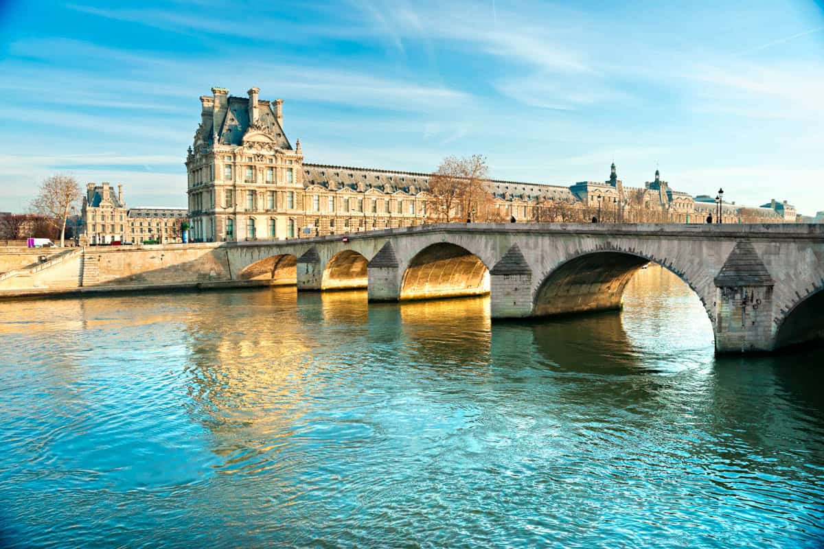 Louvre Museum in Paris, France, with Louvre riverfront wing behind an arched bridge and turquoise Seine.