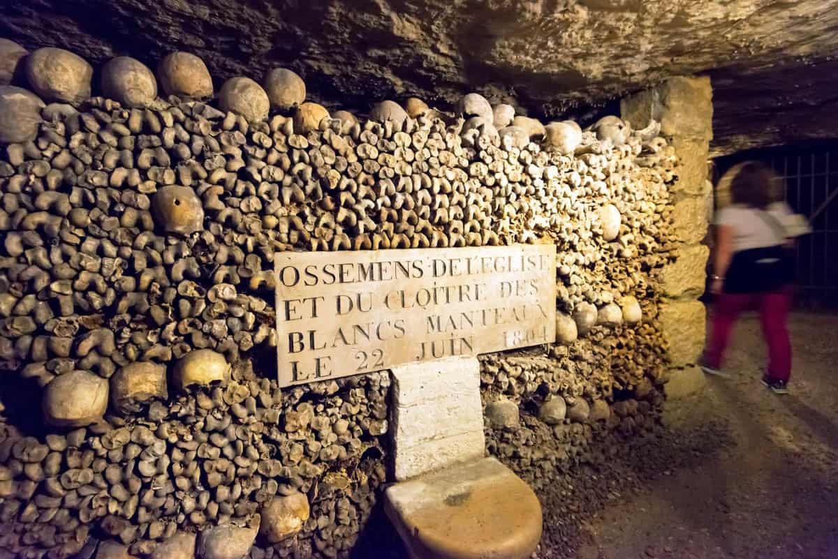 Paris Catacombs in Paris, France, with stone memorial plaque among stacked bones with a visitor passing.
