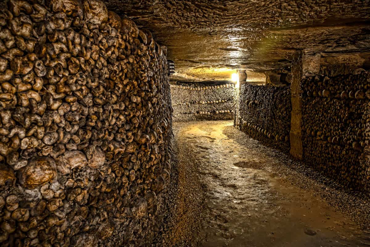 Paris Catacombs in Paris, France, with illuminated ossuary chamber with bone-lined pillars and a gravel path.