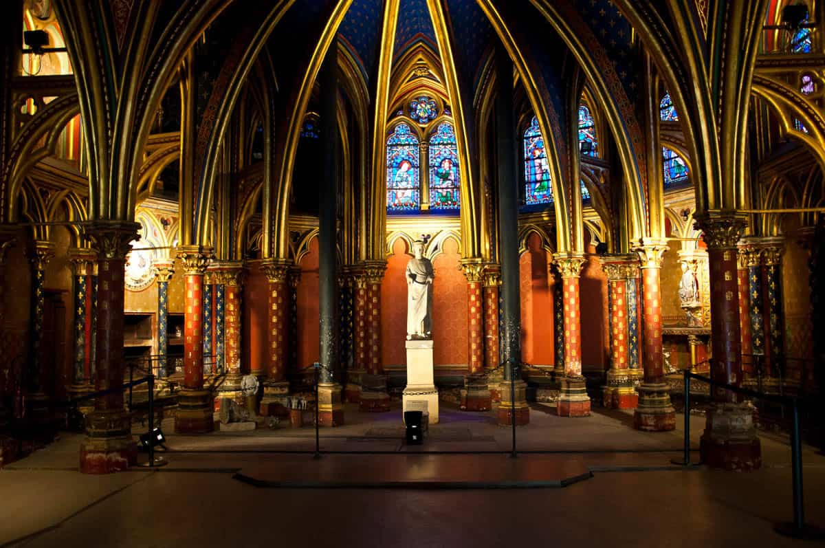 Dim lower-chapel aisle with an altar and richly painted surfaces framed by stained glass and gothic chapel at Sainte-Chapelle in Paris, France.