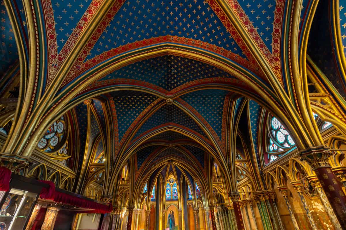 Decorated side aisle with pointed arches, columns, and stained glass with gothic chapel details around Sainte-Chapelle in Paris, France.