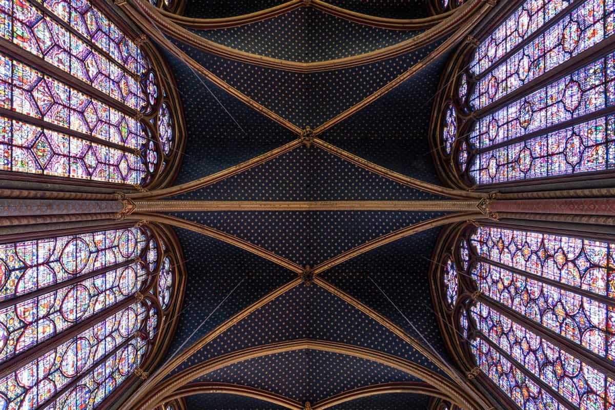 Dark blue vaulted ceiling framed by tall stained-glass windows with gothic chapel details around Sainte-Chapelle in Paris, France.
