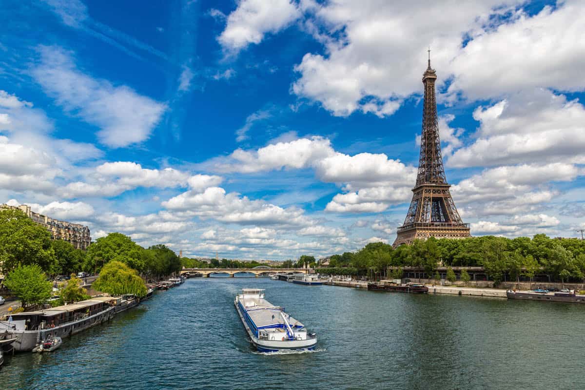 Seine River in Paris, France pairs excursion boat traveling toward the Eiffel Tower between green riverbanks with river details.