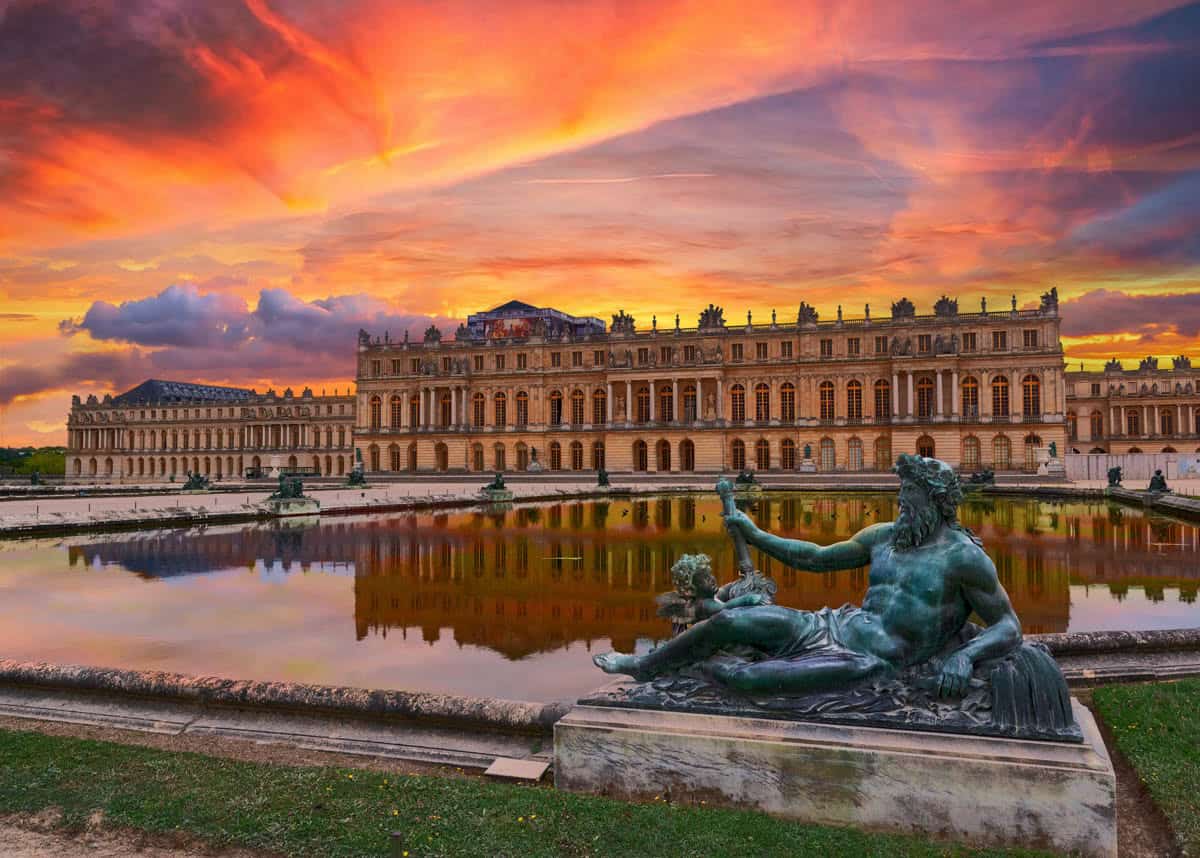 Palace and fountain sculpture reflected beneath a fiery sunset framed by palace and formal gardens at Palace of Versailles in Versailles, France.