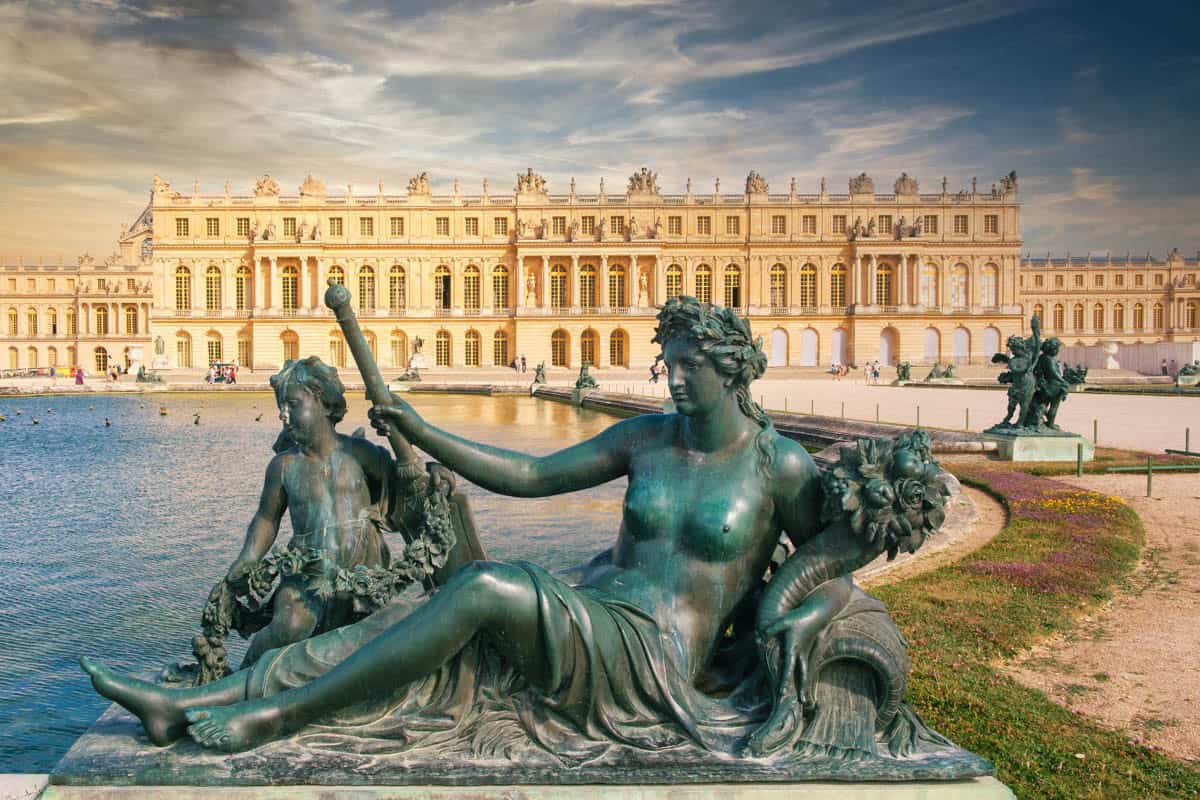 Bronze reclining figure in the foreground with the palace beyond framed by palace and formal gardens at Palace of Versailles in Versailles, France.