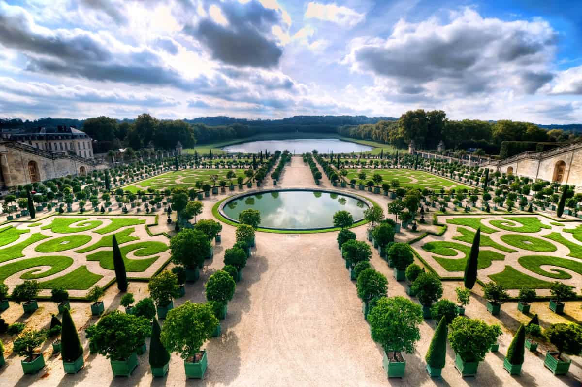 Symmetrical garden parterres and pools under dramatic clouds framed by palace and formal gardens at Palace of Versailles in Versailles, France.