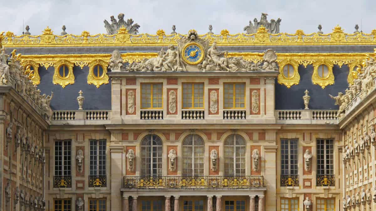 Gilded clock and sculpted ornament on the palace roofline framed by palace and formal gardens at Palace of Versailles in Versailles, France.