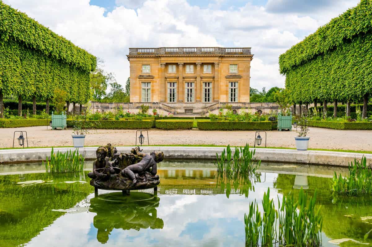 Palace of Versailles in Versailles, France, featuring garden pavilion framed by clipped hedges, lawn, pond, and sculpture beside palace features.