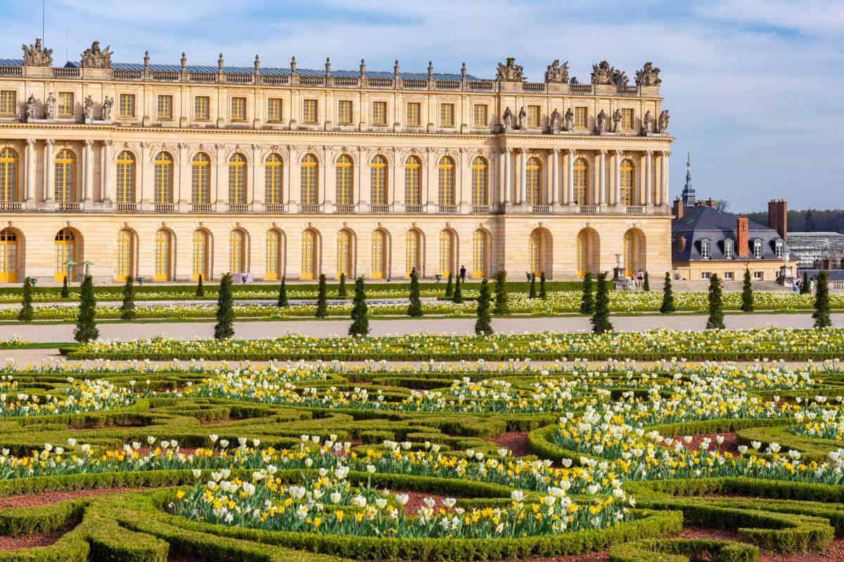 Palace of Versailles in Versailles, France, featuring palace façade behind symmetrical flowerbeds and formal lawn beside palace features.