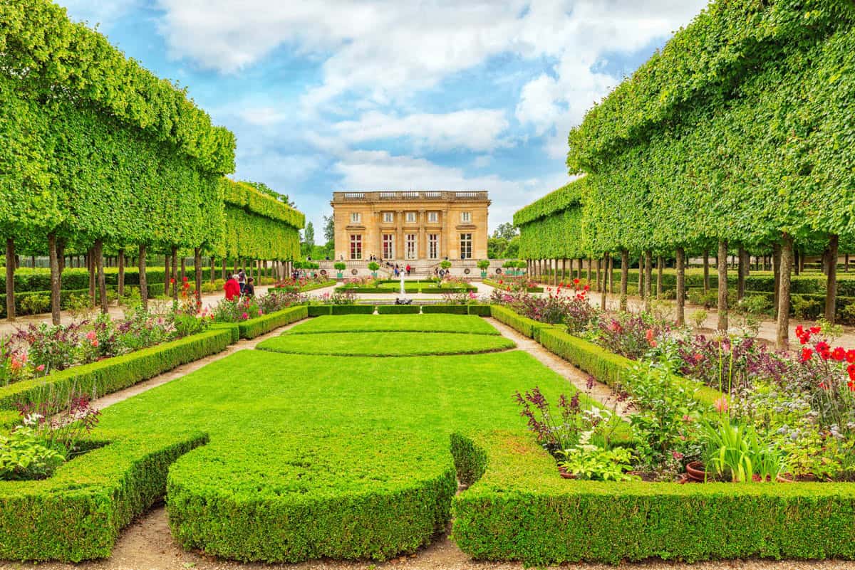 Palace of Versailles in Versailles, France, featuring tree-lined garden avenue and clipped hedges leading toward a pavilion beside palace features.