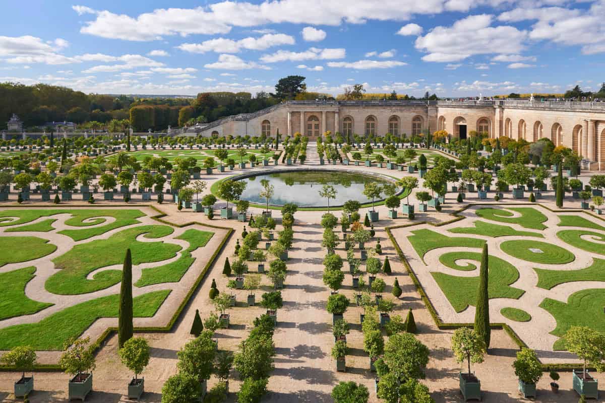 Palace of Versailles in Versailles, France, featuring geometric hedges, clipped trees, and fountains across the formal gardens beside palace features.
