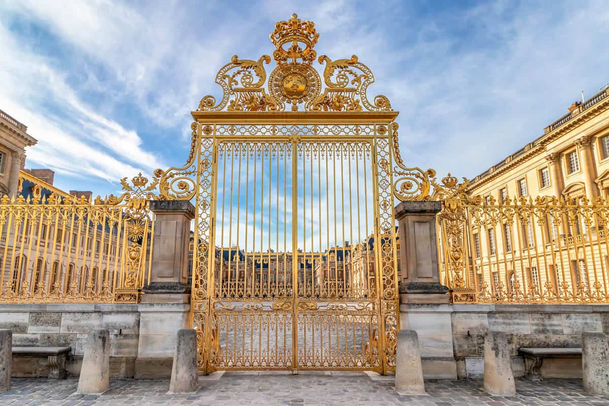 Gilded entrance gate and crest between palace wings framed by palace and formal gardens at Palace of Versailles in Versailles, France.