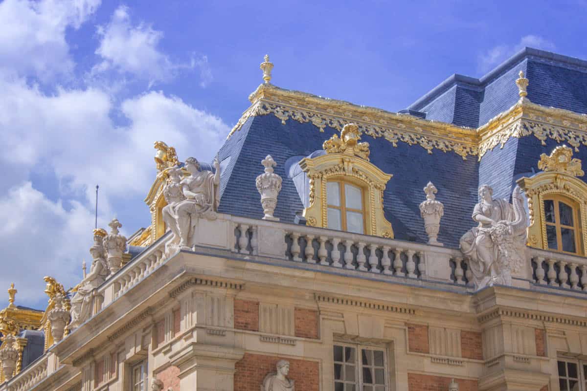 Palace of Versailles in Versailles, France, featuring slate roof, gilded cresting, and stone figures on a palace pavilion beside palace features.